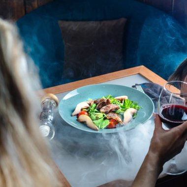 Kadın şarap içer ve taze salatayla biftek yer. Dumanlı bir nargile barında, seçici bir şekilde tabağa odaklanır.