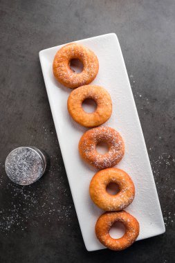 Pudra şekerli beyaz tabakta çörekler. Ev yapımı çörekler koyu taştan arka planda. Üst manzara, düz uzanma