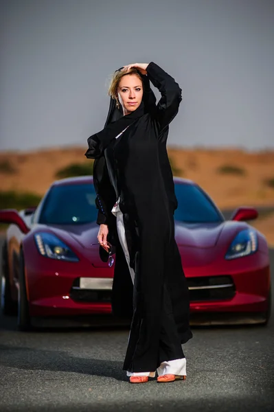 Arabic way dressed yang woman posing in red car in desert. — Stock Photo, Image