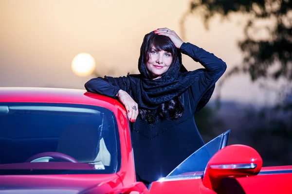 Arabic way dressed yang woman posing in red car in desert. — Stock Photo, Image