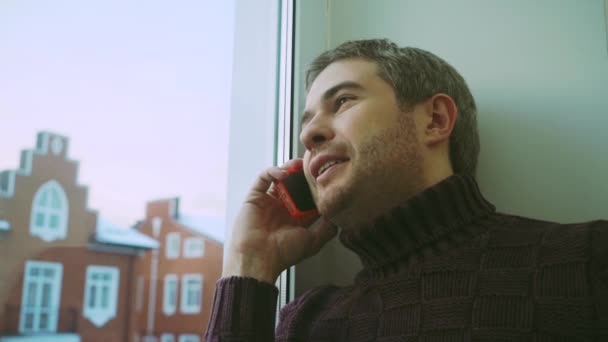 Pohledný muž ve svetru, který mluví na telefonu u okna do městských domů — Stock video