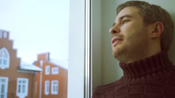 Человек в свитере подходит к окну, смотрит сквозь него и улыбается — стоковое видео