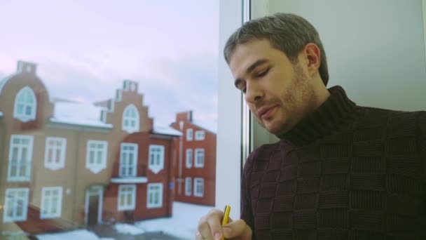 Junger Mann mit grauen Haaren schreibt in seinen Planer am Fenster — Stockvideo