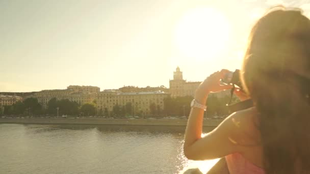 Menina morena bonita em vestido rosa tirando fotos do pôr do sol da ponte. Lento movimento clipe steadicam — Vídeo de Stock