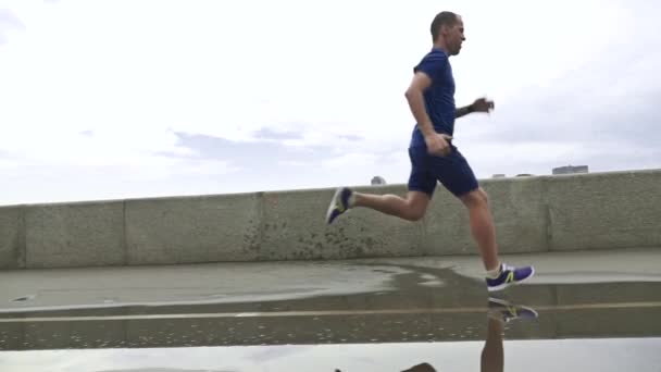 Deportista corriendo en el terraplén del río después de la lluvia y entrando en el charco. Super cámara lenta steadicam disparo a 240 fps — Vídeo de stock