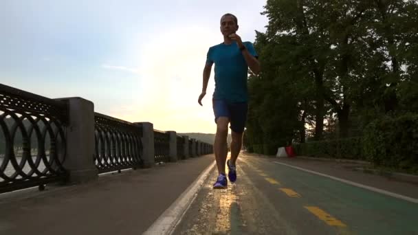 Coureur en vêtements bleus courant à la caméra le long du talus du coucher du soleil d'été de la rivière Moscou. Stéadicam au ralenti à 120 ips — Video