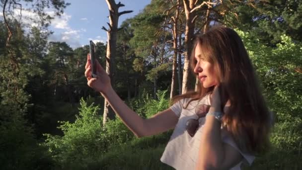 Hermosa chica morena de pelo largo haciendo selfie con su teléfono en el atardecer bosque de coníferas — Vídeo de stock