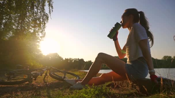Стройная девушка-велосипедистка с хвостиком пьет из бутылки. Медленный выстрел против солнца — стоковое видео
