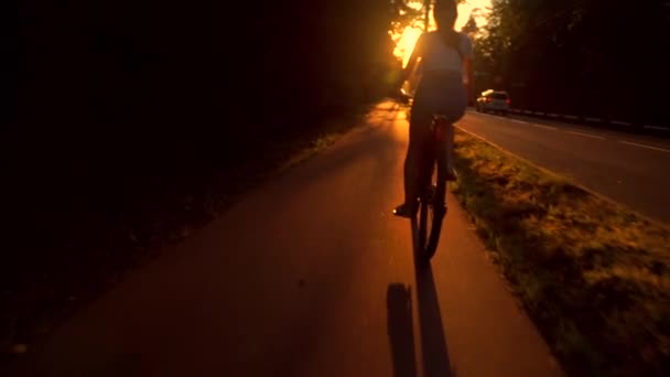 Chica morena delgada montando una bicicleta a lo largo de la carretera del atardecer verano. Disparo de seguimiento en cámara lenta — Vídeos de Stock