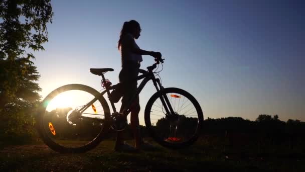 Silhuett av flicka cyklist med hästsvans frisyr promenader med sin cykel. Slow motion sköt mot solnedgången — Stockvideo