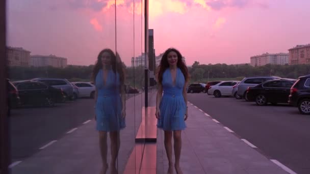 Mince fille brune en robe bleue marche dans la rue contre le coucher de soleil rose ciel, vidéo 4K — Video