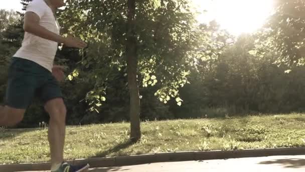 Läufer in weißem T-Shirt läuft im Park gegen pralle Sonne und Bäume. Zeitlupe Dolly Schuss, gebleichte Farben — Stockvideo