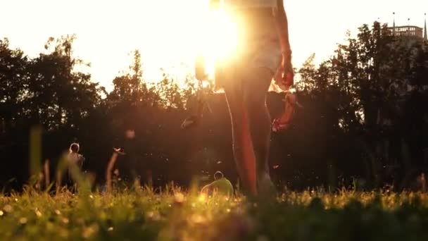 Fille mince marchant pieds nus sur l'herbe tenant ses chaussures à talons hauts à la main, tir 4K — Video