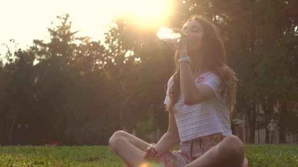 Красивая брюнетка, сидящая и пьющая воду в парке против палящего солнца, видео в 4К — стоковое видео