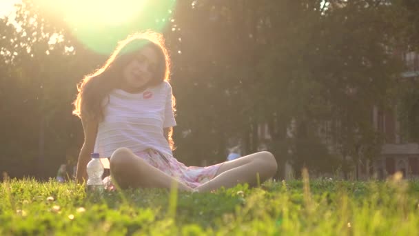 Девушка-брюнетка, сидящая и отдыхающая на траве в парке на фоне яркого солнца, снята в 4K — стоковое видео