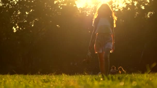 Mujer joven caminando descalza sobre la hierba sosteniendo sus zapatos de tacón alto en la mano, 4K zoom hacia fuera tiro. Colores cálidos del atardecer del verano — Vídeos de Stock