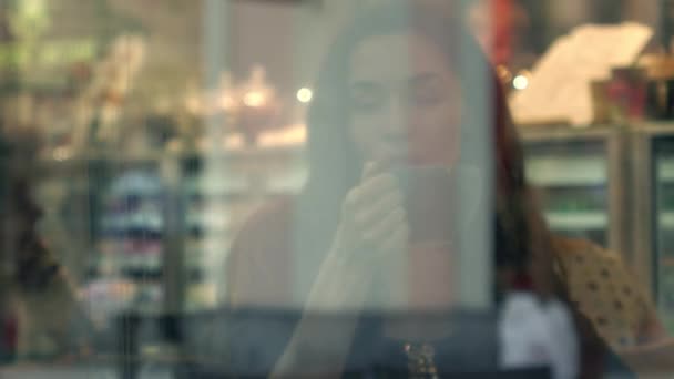 Piękna dziewczyna brunetka, pijąc gorącą czekoladę w małej kawiarni. Zobacz za pomocą okna z zewnątrz. wideo 4k — Wideo stockowe