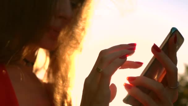 Hermosa chica con esmalte de uñas rojo usando su teléfono inteligente en el parque, colores cálidos de la puesta del sol. Vídeo en cámara lenta — Vídeo de stock