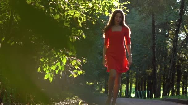 Mooie Kaukasische brunette meisje in de rode jurk wandelen langs zonnige park steegje. Slow motion video — Stockvideo