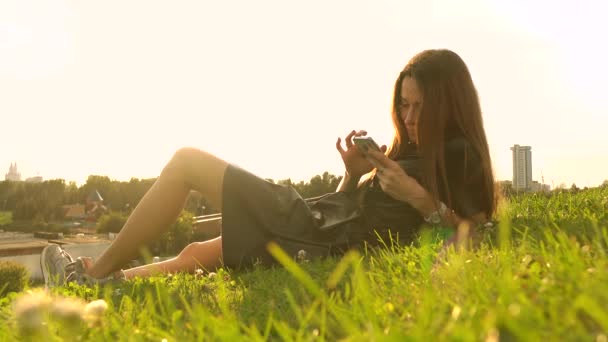 Mooie brunette meisje in zwarte jurk met behulp van haar smartphone op het gazon. Vrijetijd. 4k video — Stockvideo