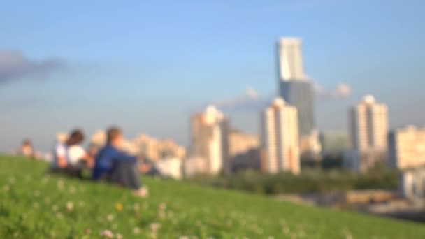 Nie do poznania młodych ludzi siedzi na trawie w parku miejskim przeciw urbanscape. 4 k bokeh tło strzał — Wideo stockowe