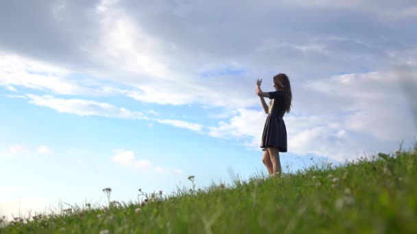 Hermosa chica morena en vestido negro de pie en la colina cubierta de hierba y hacer fotos con su teléfono. Vídeo 4K — Vídeos de Stock