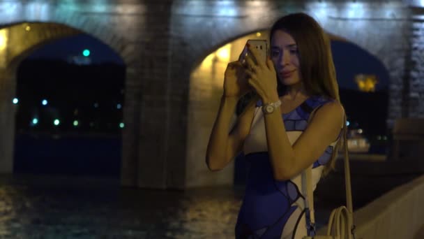 Bella ragazza bruna in abito in piedi su argine urbano sera e fare foto con il suo telefono cellulare. Video 4K — Video Stock