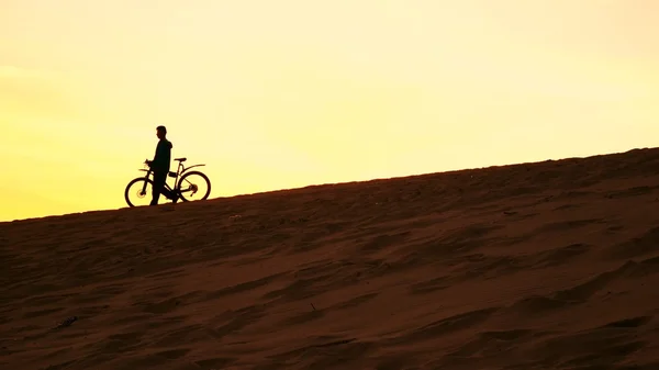 一个男人与他的自行车走在沙丘上，黄色的夕阳颜色的轮廓 — 图库照片
