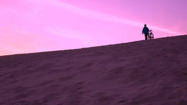 他的自行车走在沙丘上，紫色的晚霞颜色的人 — 图库照片