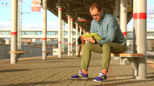 Kopasz nerdy ember, szemüveg, Vasutállomás ül, és a tábla-PC használata a naplemente. shot 4k — Stock videók