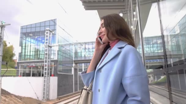 Chica feliz con abrigo azul hablando en su teléfono móvil en la estación de tren. Disparo 4K — Vídeos de Stock