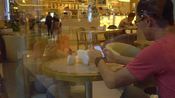 Un par en un café detrás de una gran ventana. Hombre y mujer joven hablando y utilizando sus teléfonos móviles. Disparo 4K — Vídeo de stock