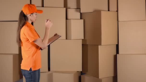 Красивая работница склада в оранжевой униформе считает предметы и делает записи против коричневых коробок. 4K студийное освещение — стоковое видео