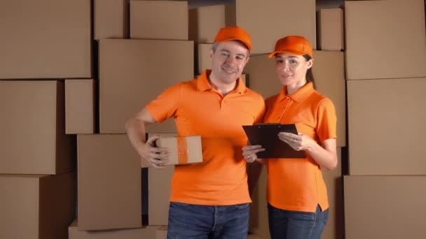 Mensajeros en uniforme naranja de pie contra las pilas de cartón marrón en el fondo. Personal de la empresa de entrega. 4K video de estudio — Vídeo de stock