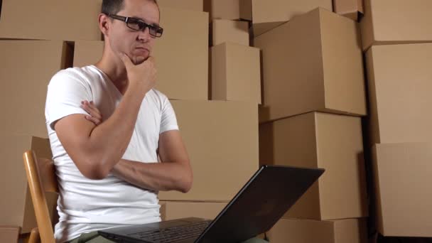 Техника маленького интернет-магазина за работой. Человек в очках думает и печатает на ноутбуке против стопок коробок. 4K клип — стоковое видео