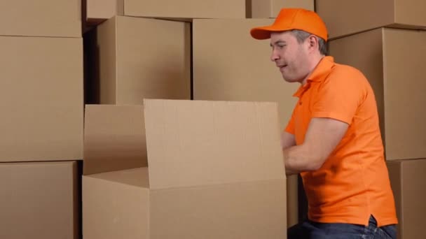 Warehouse worker in orange uniform packaging big carton with duct tape gun. Multiple boxes background. 4K video — Stock Video