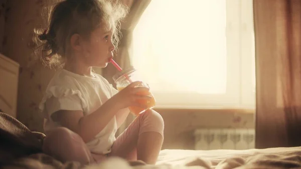 Menina loira bonito bebe suco de laranja com uma palha em casa — Fotografia de Stock