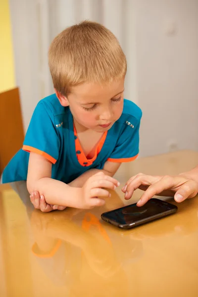 Niño juega con un teléfono inteligente en la mesa de la cocina — Foto de Stock
