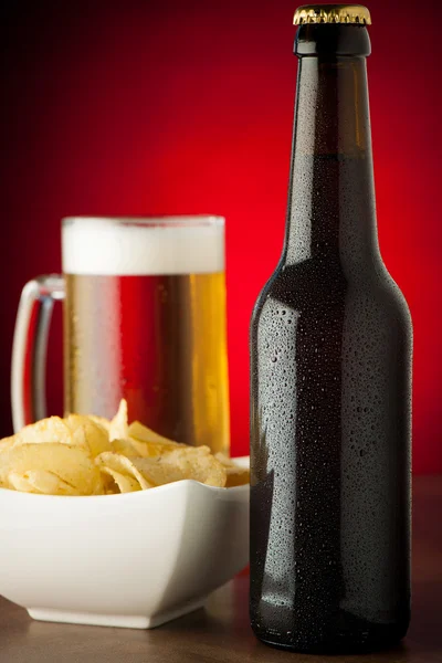 Bottle, glass of beer and potatoe chips on stone table over red — Stock Photo, Image