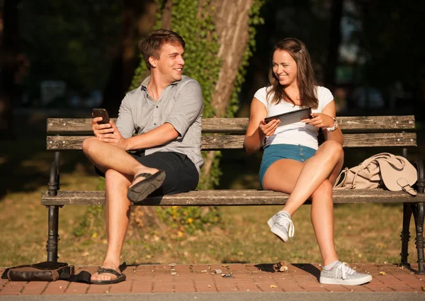 Pareja joven divirtiéndose en un banco en el parque mientras socializa ove — Foto de Stock