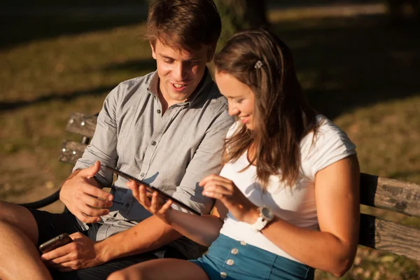 Jovem casal se divertindo em um banco no parque enquanto socializa ove — Fotografia de Stock