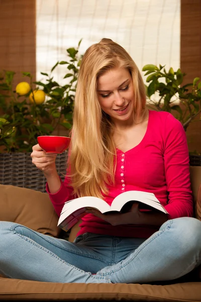 Frau sitzt gemütlich auf Sofa, liest Buch und trinkt Tee — Stockfoto