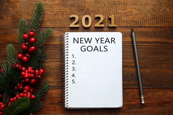 New Year goal list 2021. Christmas tree, red berries, pencil and notebook for writing goals or results of the year. Top view. Flat lay on wooden background. Festive, holiday and motivation concept.