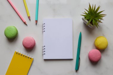 Not defterleri, kalemler, kalem, bitki ve mermer zemin üzerinde badem kurabiyeleri olan çalışma alanı. Üst manzara, düz yatış. Eğitim ve iş konsepti. Modayı takip eden renkli kadın işyeri.