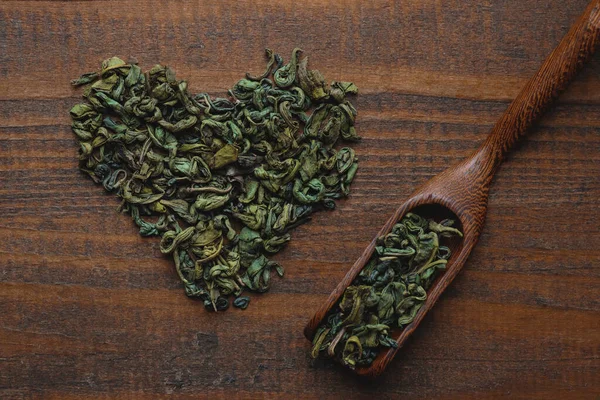 Dried green tea leaves in the shape of heart and spoon on a wooden background. Selective focus, top view, flat lay.