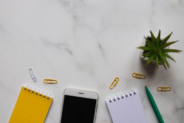 Üst kat ofis masası. Not defterleri, kalemleri, akıllı telefonları ve açık gri mermer arka planlı sulu bitkileri olan modern çalışma alanı. Düz yatıyordu.