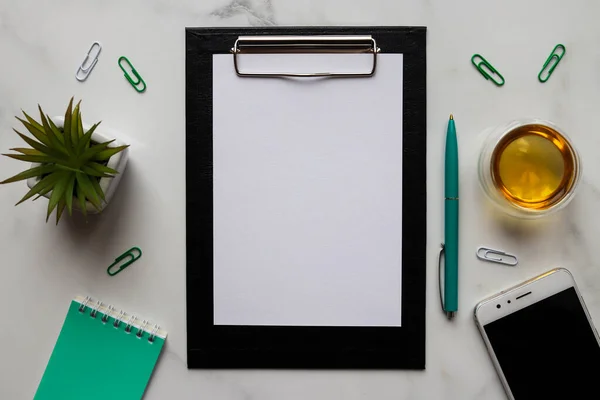 Workplace with clipboard with empty sheet of paper, pen, smartphone, cup of tea and succulent plant on marble background. Office table desk. Flat lay, top view