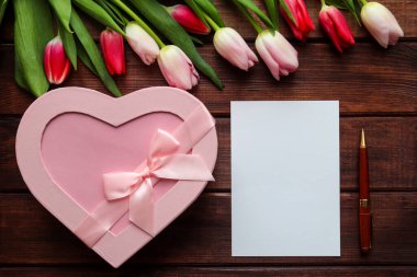 Koyu ahşap zemin üzerinde bir buket lale, kalp şeklinde hediye kutusu, içi boş kağıt ve kalem. Kutlamalar için şenlikli bir maket. Uzayı metin için kopyala. Yakın plan..