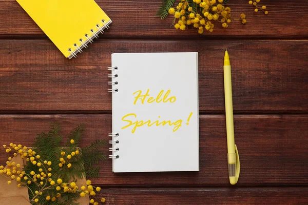Springtime concept. Notepad with the inscription Hello Spring on a wooden background and yellow mimosa flowers decor. Top view, flat lay.