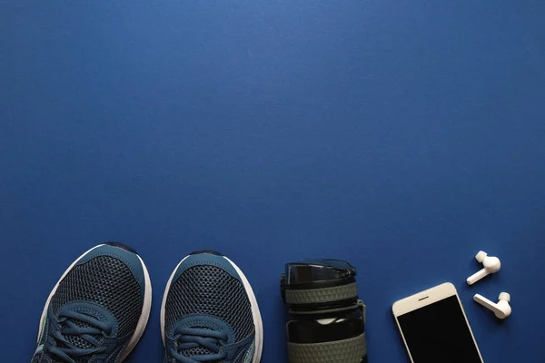 Sağlıklı yaşam tarzı, spor anlayışı. Spor malzemeleri, spor ayakkabıları, su şişesi, akıllı telefon ve fotokopi alanı olan koyu mavi arka planda kulaklık. Düz yatış, üst görünüm